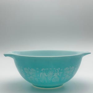 May include: A turquoise milk glass mixing bowl featuring a white printed pattern of figures, wheat sheaves, and farm-related motifs. The bowl has two integrated handles on opposite sides and a smooth, rounded base, showcasing a classic design.