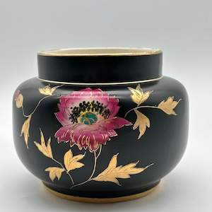Peut inclure: Un vase en céramique noir mat avec un corps large et arrondi et un col court. L'extérieur présente une fleur peinte à la main rose et blanche avec un centre sombre, entourée de motifs de feuillage en feuille d'or et un bord doré à la base.