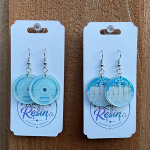 May include: Two pairs of circular dangle earrings on white display cards. The left pair features a light blue vinyl record design. The right pair displays a group of five figures in white clothing against a blue background. Both have silver hooks.