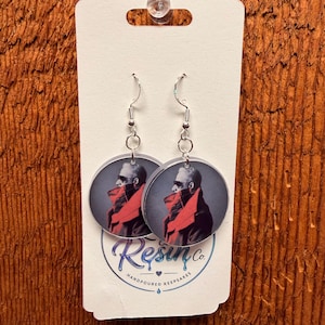 May include: A pair of round dangle earrings featuring a photographic print of a person wearing sunglasses and a red scarf. The earrings hang from silver-toned metal hooks on a white display card with the text Resin Co. Handpoured Keepsakes.