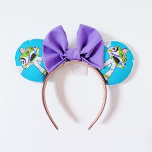 May include: A headband featuring two round ears covered in a light blue fabric with a repeating pattern of Buzz Lightyear characters against a white cloud background. A large, solid purple fabric bow is centered between the two ears.