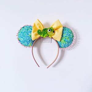 Peut inclure: Un serre-tête avec deux oreilles circulaires recouvertes d'un motif floral bleu et vert avec une bordure à pompons roses. Un grand nœud en tissu jaune est placé entre les oreilles, centré avec une figurine de caméléon vert aux grands yeux.