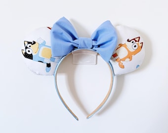 May include: A headband featuring two round, padded ears covered in white fabric with a repeating Bluey character print. A large, solid light blue fabric bow is centered between the ears. The headband frame is light blue with a tan inner lining.