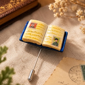 Peut inclure: Une épinglette représentant un livre miniature ouvert aux pages jaunies, posé sur une petite base rectangulaire bleue. Les pages du livre présentent deux petits timbres-poste illustrés de personnages. Une fine épingle en métal argenté dépasse.