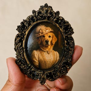 May include: A small oval portrait of a golden retriever wearing a floral hat and a high-collared cream dress, encased in an ornate, black and gold-toned frame with intricate scrollwork and decorative relief detailing along the edges.