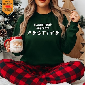 May include: A dark green long-sleeve sweatshirt featuring the white text Could I BE any more FESTIVE with colorful dots between the letters. The sweatshirt is paired with red and black buffalo plaid pants and a white ceramic Santa Claus mug.
