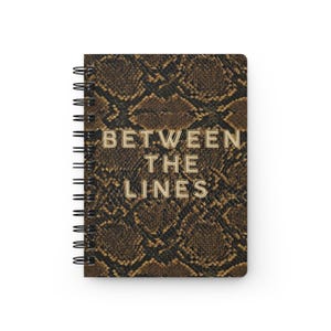 May include: A spiral-bound notebook featuring a brown and black snakeskin pattern on the cover. The text "BETWEEN THE LINES" is printed in a light beige, block-style font across the centre of the front cover, aligned vertically in three rows.