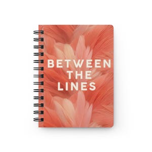 May include: A spiral-bound notebook featuring a cover with a soft coral and peach-toned feather pattern. The text "BETWEEN THE LINES" is printed in a clean, white, sans-serif font across the center of the front cover, aligned vertically.