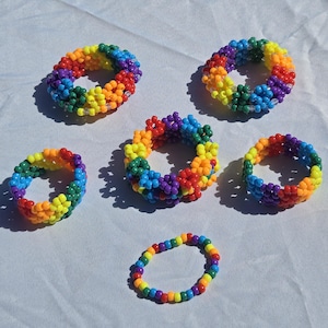 May include: A collection of six handmade bracelets crafted from small, vibrant, multi-colored plastic beads. Five of the bracelets feature a thick, textured, multi-layered design, while one bracelet is a single, thin strand of rainbow-colored beads.