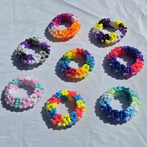 May include: Eight circular bracelets crafted from small, colorful plastic beads. The bracelets feature various patterns, including solid rainbow, pink and white, blue and green, purple and black, and multi-colored combinations arranged in a repeating grid.