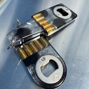 May include: Two bottle openers made of black and white marbled resin, each featuring a row of embedded brass-colored bullet casings. The openers have a circular metal head secured with two screws and a metal keyring attached to the top end.