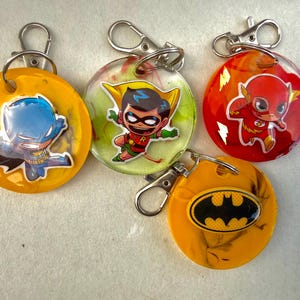Peut inclure: Quatre porte-clés circulaires en résine avec des fermoirs mousquetons en métal. Un porte-clés jaune présente une figurine de Batman bleue, un vert translucide présente Robin, un rouge présente Flash et un jaune présente un logo de chauve-souris noir.