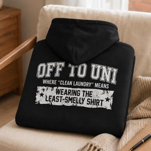 Op de afbeelding: Een zwarte hoodie met witte tekst in een verweerde stijl die luidt OFF TO UNI WHERE CLEAN LAUNDRY MEANS WEARING THE LEAST-SMELLY SHIRT. Het ontwerp bevat twee kleine sterren die de onderste tekstregel op het kledingstuk flankeren.