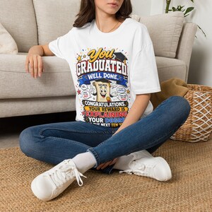 May include: A white short-sleeved t-shirt featuring a colorful graphic with the text "You Graduated Well Done. Congratulations. Your reward is explaining your degree for the next ten years." The design includes a cartoon diploma with a graduation cap.