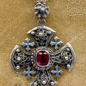 May include: A silver-toned metal cross pendant featuring an intricate filigree design with small silver spheres and a central rectangular red gemstone. The pendant is suspended from a matching silver-toned metal chain link necklace.