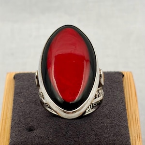 May include: A silver ring featuring a large, oval-shaped cabochon stone with a vibrant red center surrounded by a deep black border. The metal band displays intricate, openwork filigree detailing along the sides, providing a decorative texture.