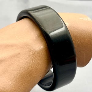 Może przedstawiać: Jednolita czarna bransoletka typu bangle o gładkim, polerowanym i błyszczącym wykończeniu. Bransoletka ma szeroki, zaokrąglony kształt o stałej grubości, prezentując głęboki, jednolity czarny kolor oraz odblaskową powierzchnię.
