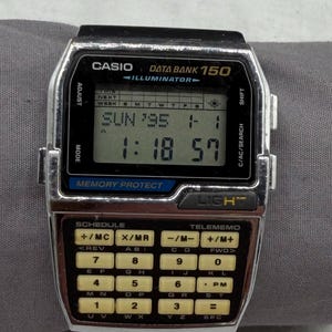 May include: A silver and black Casio Data Bank 150 Illuminator digital watch featuring a rectangular LCD screen displaying time and date, and a numeric keypad below. The watch includes buttons labeled Adjust, Shift, Mode, and C/AC/Search.