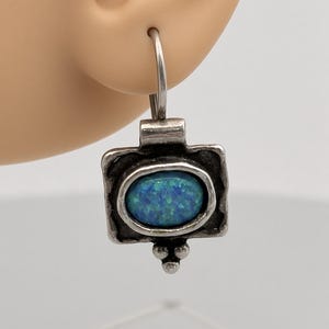 May include: A silver-toned earring featuring a rectangular frame with a dark, oxidized finish. Centered within the frame is an oval-shaped blue opal cabochon with iridescent flecks. The earring hangs from a curved wire hook and has three small silver spheres at the bottom edge.