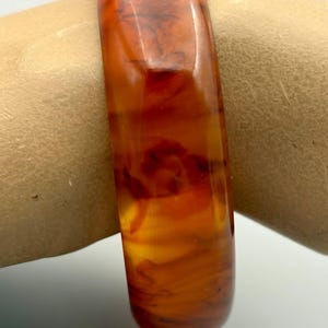 May include: A smooth, polished bangle bracelet featuring a translucent amber-colored resin material with deep, swirling dark brown and reddish-orange marbled patterns throughout the rounded, thick band structure, creating a glossy, multi-tonal appearance.