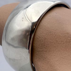 May include: A wide, polished silver-tone bangle bracelet with a smooth, reflective surface and a slightly irregular, hammered-style contour. The bracelet features a thick, rounded band design that maintains a consistent, high-shine metallic finish.