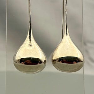 May include: A pair of polished silver-tone drop earrings featuring a smooth, rounded teardrop shape. Each earring hangs from a curved wire hook, showcasing a reflective, metallic surface that catches light with a clean, minimalist aesthetic.
