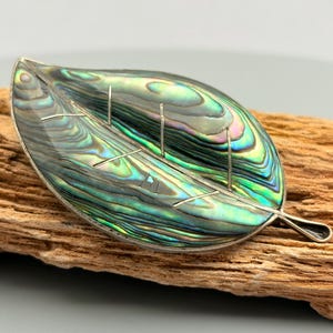 Könnte beinhalten: Eine blattförmige Brosche mit einer Einlage aus poliertem Abalone-Muschelgehäuse, das in schillernden Grün-, Blau- und Violetttönen schimmert. Das Stück ist von einem dünnen silberfarbenen Metallrand mit feinen silberfarbenen Adern umgeben.