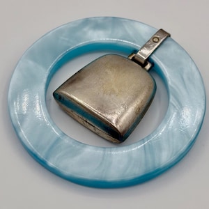 May include: A baby rattle featuring a smooth, circular light blue marbled ring made of a plastic-like material. Attached to the ring is a metallic, silver-toned bell with a slightly tarnished finish, secured by a small metal loop and rivet.