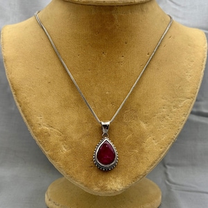May include: A silver necklace featuring a teardrop-shaped pendant with a deep red stone. The pendant is framed by a beaded silver border and hangs from a thin, flexible silver chain. The design is simple, focusing on the contrast between the metallic silver and the dark red gemstone.