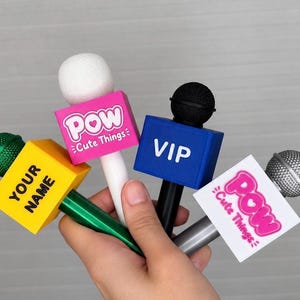 May include: Four handheld microphone flags in yellow, pink, blue, and white. The yellow flag says YOUR NAME, the pink and white flags say Pow Cute Things, and the blue flag says VIP. Each features a foam or metallic microphone head.