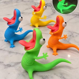 May include: Four lizard-shaped fidget toys in blue, yellow, orange, and green. Each figure features a wide-open mouth with a pink tongue, large white eyes with black pupils, and a ribbed torso. An inset shows the green lizard glowing in the dark.
