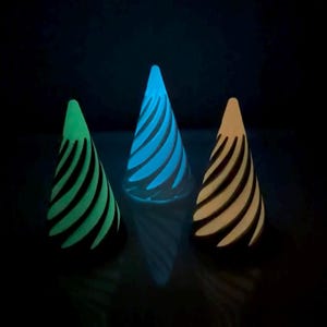 May include: Three cone-shaped objects with a spiral cutout pattern arranged in a row. The left cone glows green, the center cone glows blue, and the right cone glows orange. Each object emits a soft light from within against a dark background.