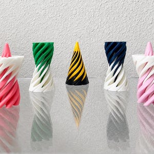 May include: Five 3D printed objects arranged in a row. Two outer objects are white with pink spiral patterns. Two inner objects are white with green or dark blue spiral patterns. The center object is a solid cone with black and yellow spiral stripes.
