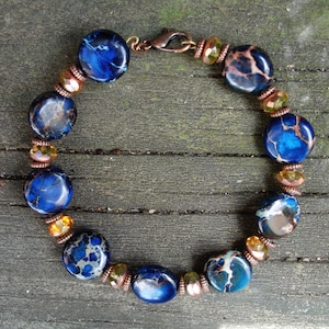 May include: A handcrafted bracelet featuring large, round, blue jasper beads with brown and white veining. The bracelet also includes smaller, amber-colored beads and copper-toned spacers, secured with a copper clasp.