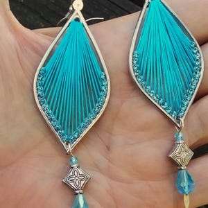 May include: A pair of turquoise thread earrings with a silver-colored frame and accents. The earrings have a teardrop shape with a beaded border and a dangling silver charm with a blue bead.