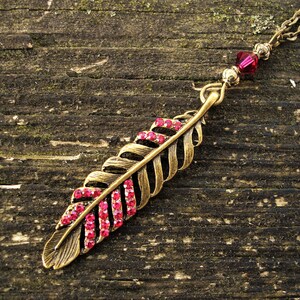 May include: A vintage-style necklace featuring a feather pendant. The pendant is gold-toned with a row of pink gemstones. The necklace has a delicate chain and a small red bead near the clasp. The background is a weathered wooden surface.