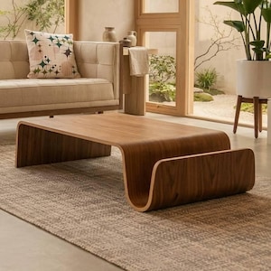 Peut inclure: Une table basse à profil bas fabriquée en bois courbé avec un grain brun riche. La table présente un design continu et incurvé qui crée un compartiment de rangement intégré sur un côté, reposant directement sur un tapis texturé aux tons neutres.
