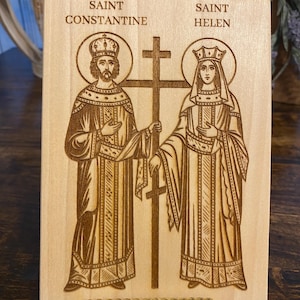 May include: A rectangular light-colored wooden plaque featuring a laser-engraved illustration of Saint Constantine and Saint Helen standing on either side of a tall cross. The text SAINT CONSTANTINE and SAINT HELEN is engraved above each figure.