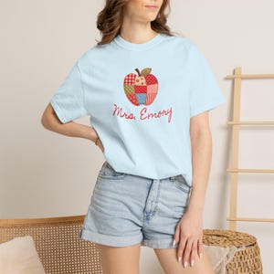 Puede incluir: Una camiseta de manga corta de color azul claro con un gráfico central de una manzana estilo patchwork en patrones rojos, blancos y verdes. Debajo de la manzana, el texto Mrs. Emory está impreso en una fuente cursiva roja en el frente.
