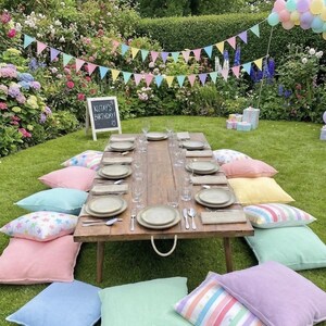 May include: A low wooden picnic table set on grass with place settings including plates, glasses, and cutlery. The table is surrounded by colourful pastel floor cushions, a string of triangular bunting, a balloon arch, and a chalkboard sign.