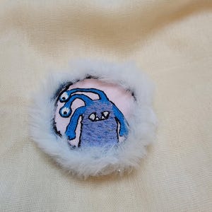 May include: A circular embroidered patch featuring a blue monster with two eyes on stalks and sharp white teeth. The central design is stitched on a light pink fabric background and is surrounded by a thick, fluffy white faux fur border.