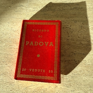 May include: A rectangular red book with gold lettering and a decorative border. The cover features the text RICORDO DI PADOVA and 20 VEDUTE 20. The book has a textured surface and is positioned at an angle, casting a distinct shadow to its right side.