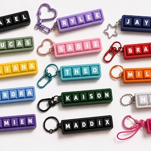May include: A collection of rectangular keychains featuring mechanical keyboard-style keys with white letters spelling names including AXEL, RYLEE, JAYDEN, LUCAS, SADIE, BRAYLON, ELIANA, THEO, MILAN, AURORA, KAISON, NOVA, DAMIEN, MADDIX, and ZOEY with metal clips.