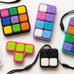 Op de afbeelding: Een verzameling sensorische siliconen fidget-tools in verschillende vormen en maten, met kleurrijke vierkante knoppen met diverse tactiele texturen zoals spiralen, stippen en strepen. Sommige items zijn voorzien van zwarte koorden.