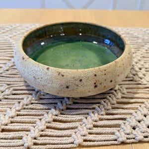 May include: A round ceramic bowl with a textured, speckled beige exterior and a smooth, glossy interior glazed in shades of deep forest green and dark brown. The bowl features a low profile and a thick, rounded rim, showcasing a handcrafted appearance.