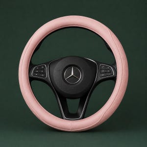Könnte beinhalten: Ein hellrosa Lenkradbezug mit texturiertem Rautenmuster, der auf einem schwarzen Mercedes-Lenkrad angebracht ist. In der Mitte des Lenkrads befindet sich das silberne Mercedes-Benz-Logo, mit integrierten schwarzen Bedientasten an den Speichen.