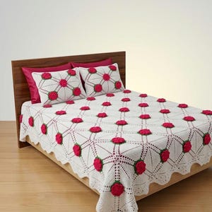 May include: A white crocheted bedspread and two matching pillow shams featuring a repeating diamond pattern with raised, three-dimensional dark pink rose motifs at each intersection. The edges of the bedspread have a scalloped crochet border.