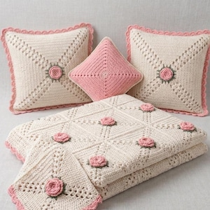 May include: A set of two square cream-colored crochet pillows with pink rose accents and scalloped edges, one small pink diamond-shaped crochet pillow, and a folded cream-colored crochet blanket featuring pink rose motifs and scalloped borders.