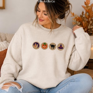 May include: A cream-colored crewneck sweatshirt featuring four circular embroidered patches across the chest. The patches display a ghost, a jack-o'-lantern, a black cat, and a purple witch hat. The text Ollie & Ella Co. appears above.