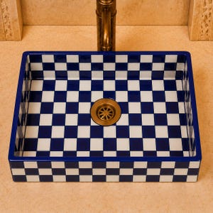 May include: A square ceramic vessel sink featuring a bold blue and white checkerboard pattern on both the interior and exterior surfaces. The basin includes a centered, circular metallic drain fixture with a bronze-toned finish and a starburst grate.